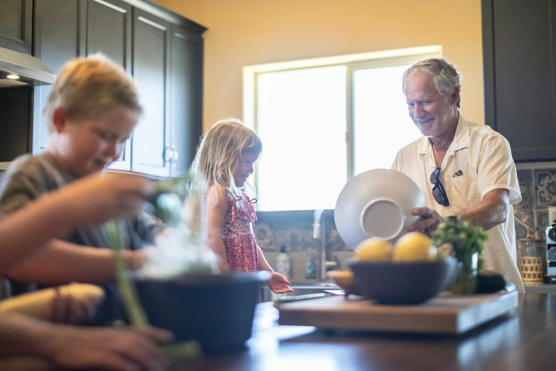 A grandfather and his grandchildren preparing food in a sunlit home kitchen, placeholder image evoking a give-back recipient family.